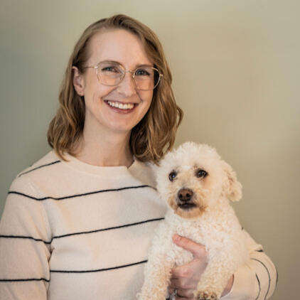 Liz, a white woman with glasses and shoulder-length blonde hair, smiles while holding an adorable small white dog named Mops.
